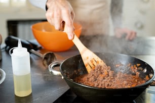 木のへらを持つ成熟した男性の手が、台所のテーブルのそばに立って料理をしながら、電気ストーブのフライパンでひき肉を混ぜる