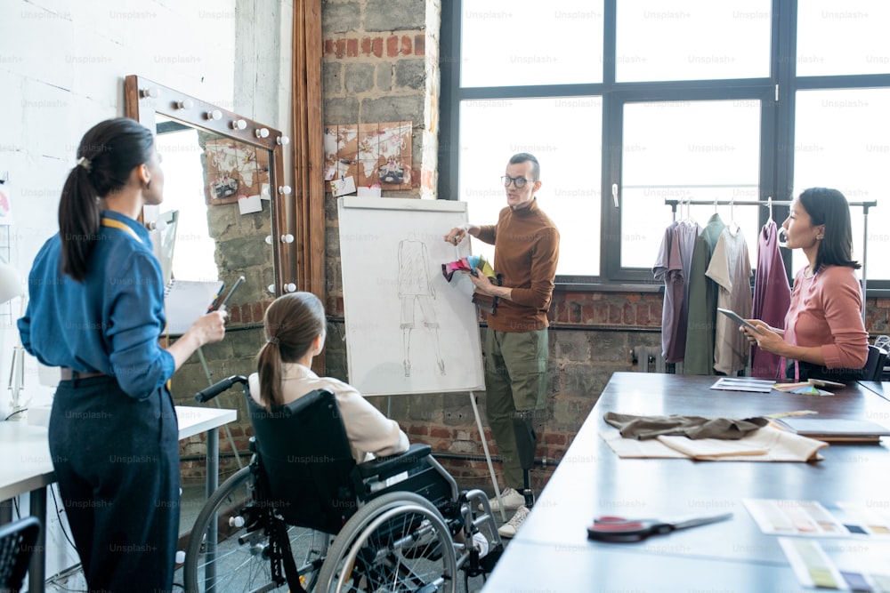 Young disable male designer with textile samples standing by whiteboard while explaining his view of new collection to colleagues at seminar