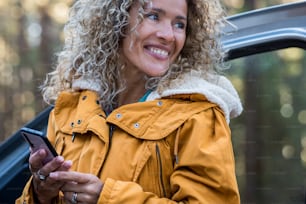 Happy cheerful beautiful young woman smile and enjoy outdoor leisure activity using phone outside her car - travel and lifestyle female people alone - blonde curly lady with yellow jacket