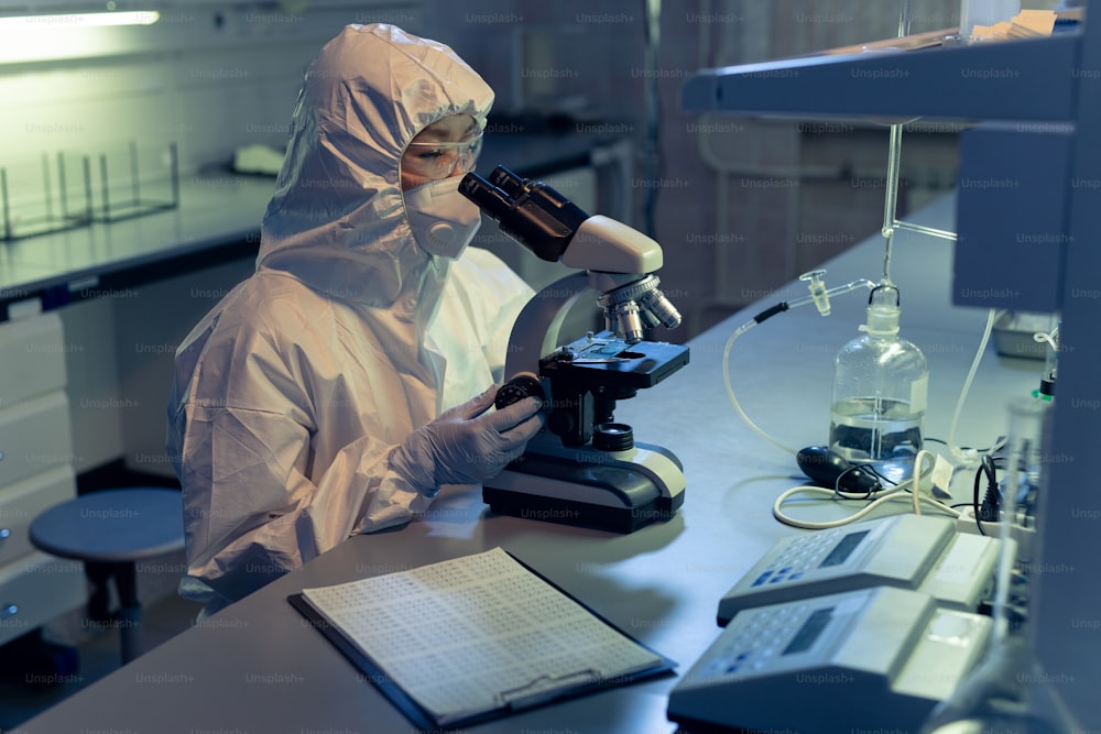 Químico em roupas de trabalho de proteção examinando amostras através do microscópio no laboratório