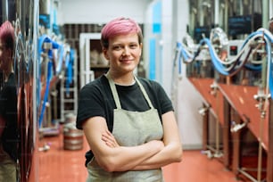 Young smiling blond female worker of beer production factory in workwear crossing arms by chest while standing against processing equipment