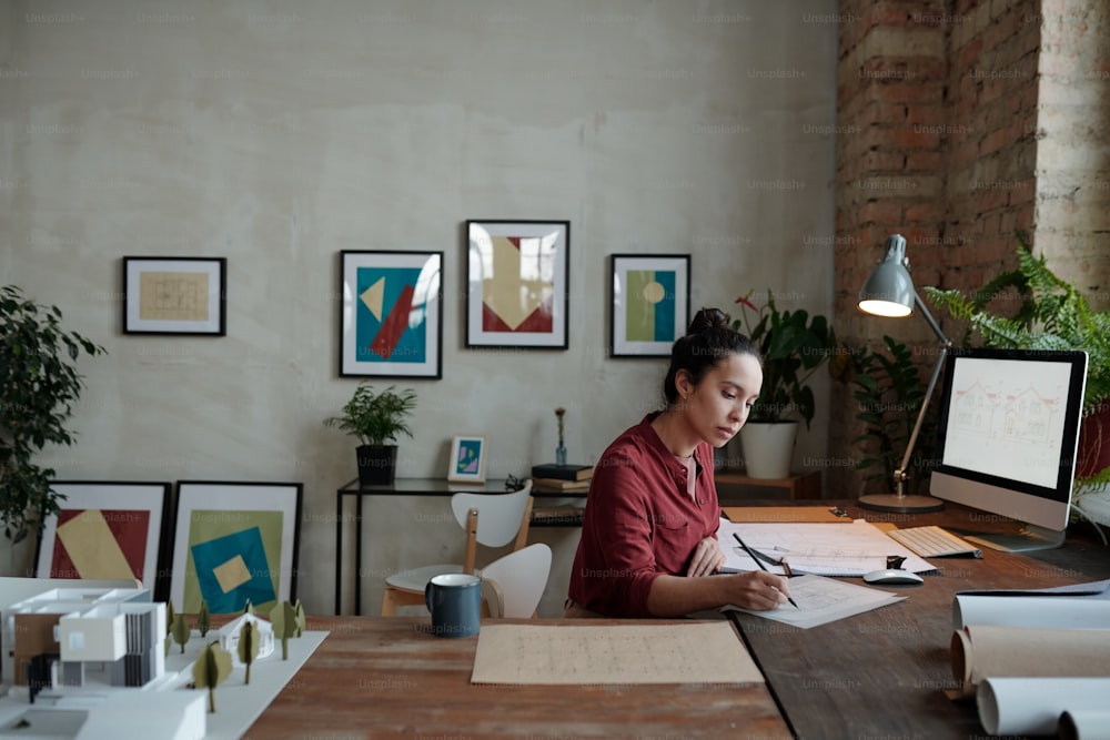 Mujer joven de raza mixta ocupada con moño de pelo sentada en el escritorio y dibujando boceto mientras trabaja en la oficina con cuadros en la pared