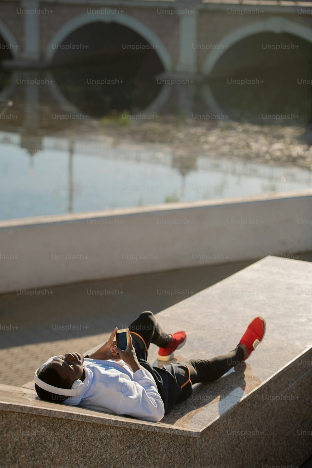Young relaxed African man in sportswear and white headphones lying by riverside after outdoor training and enjoying rest with music