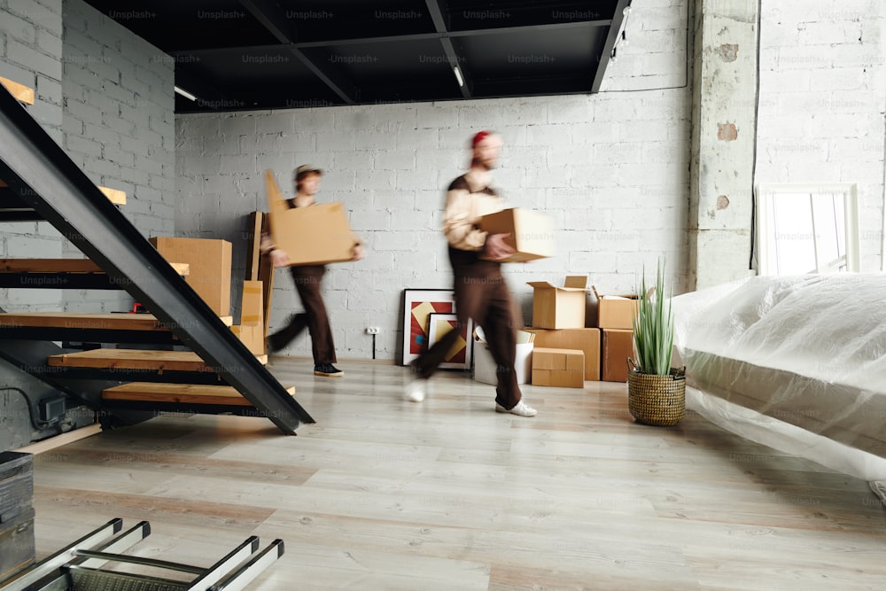 Blurred motion of two young loaders carrying packed cardboard boxes while helping to deliver packages to new flat, house or studio