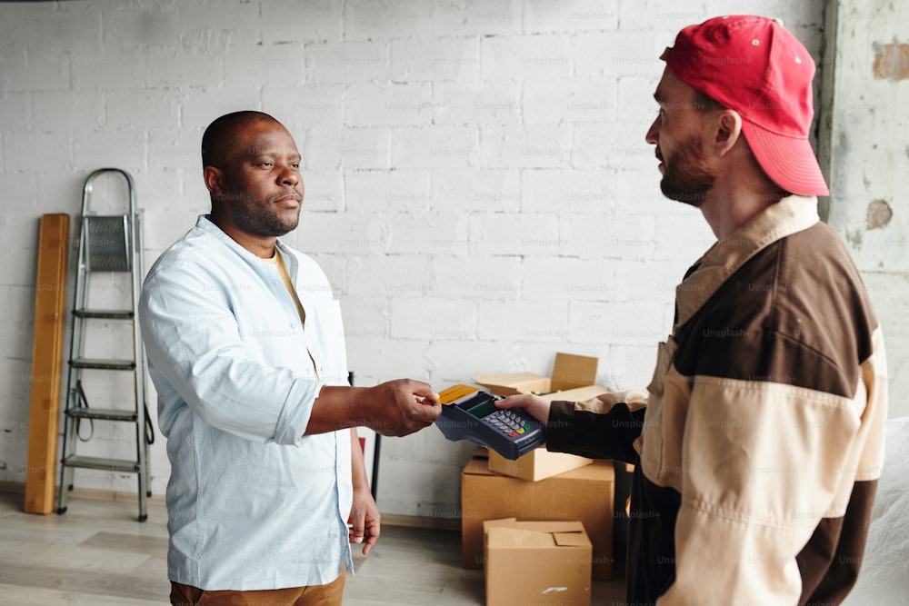 Cargador joven en terminal de retención de ropa de trabajo mientras que el cliente masculino africano paga por la carga y la prestación del servicio con tarjeta de crédito