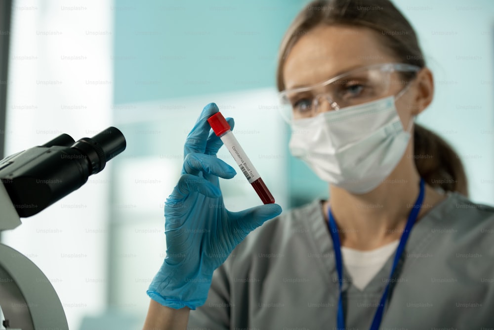 Primo piano di un giovane ricercatore di laboratorio in maschera e occhiali in piedi davanti al microscopio e con in mano un campione di sangue del paziente affetto da coronavirus