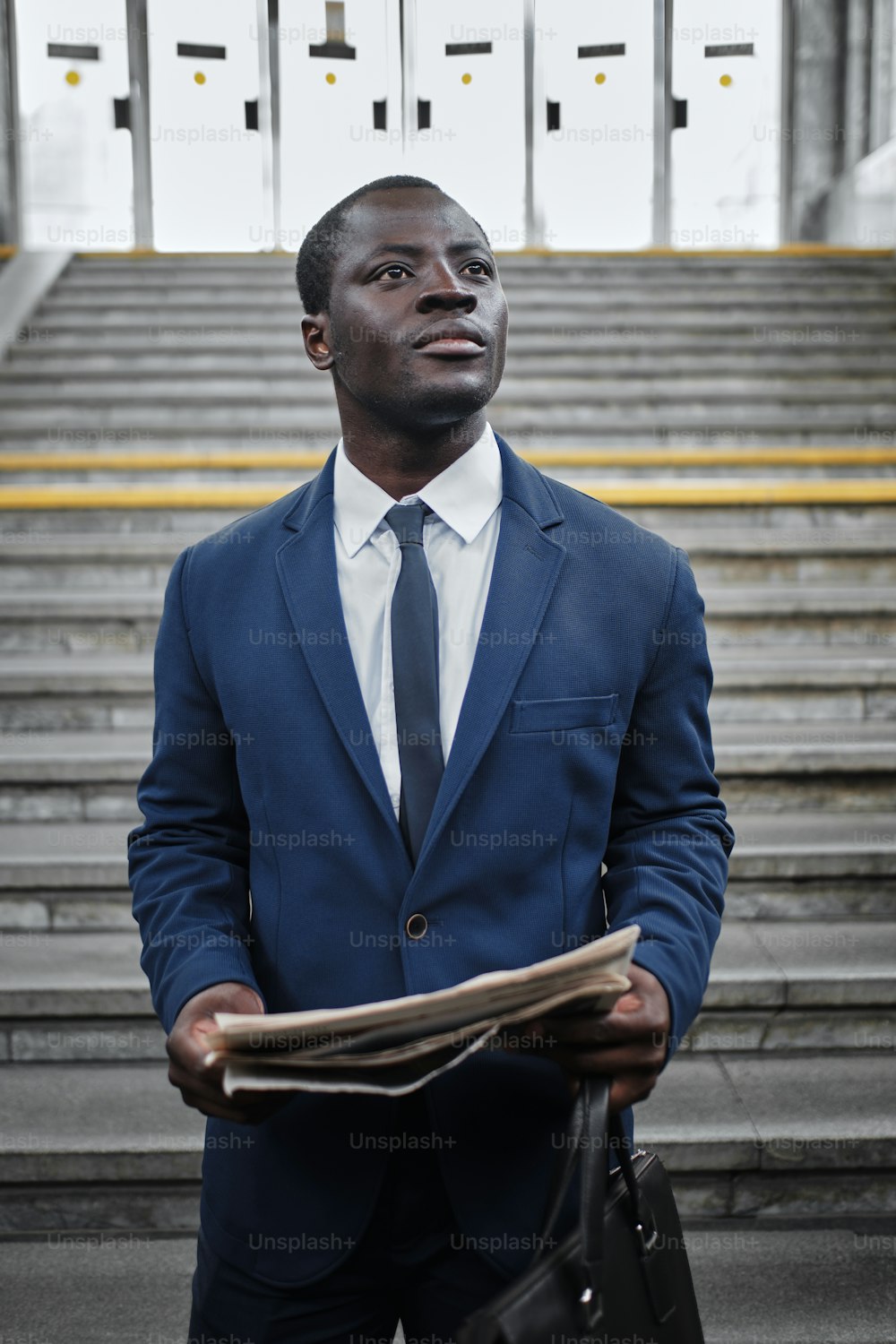 Portrait of serious confident black man wearing blue suit and tie, reading newspaper at subway station