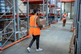 Storage workers in reflective vests moving boxes while distributing items at stockroom with metal frame shelves