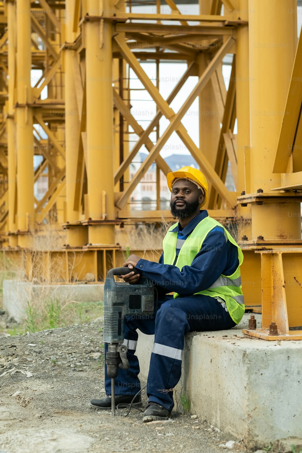Retrato de un trabajador de la construcción africano en ropa de trabajo mirando a la cámara mientras está sentado al aire libre con una herramienta de trabajo