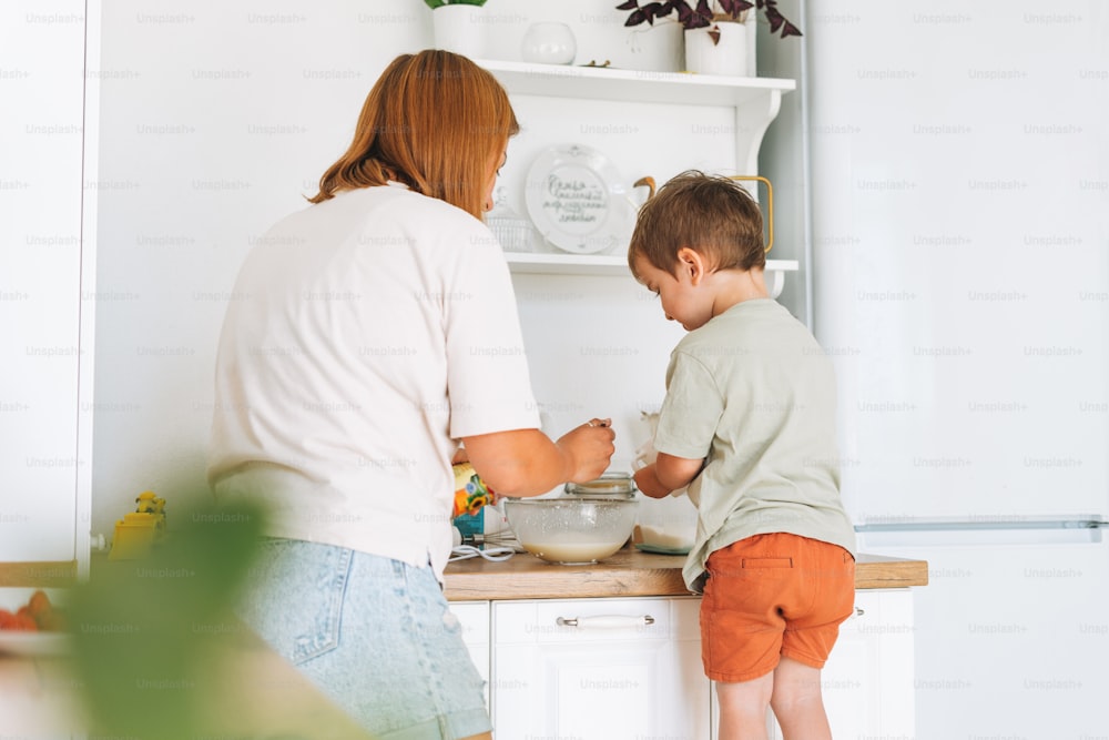 Mãe da mulher nova e seu filho menino cozindo juntos na mesa na cozinha brilhante em casa