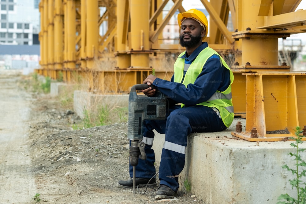 Retrato de un trabajador de la construcción africano con casco de trabajo sentado con driil al aire libre