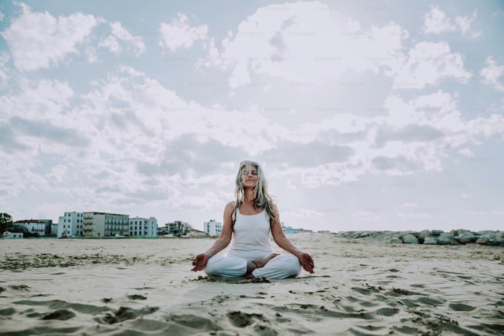 Senior woman in lotus pose sitting on the sand - Yoga at beach - Calm and meditation concept