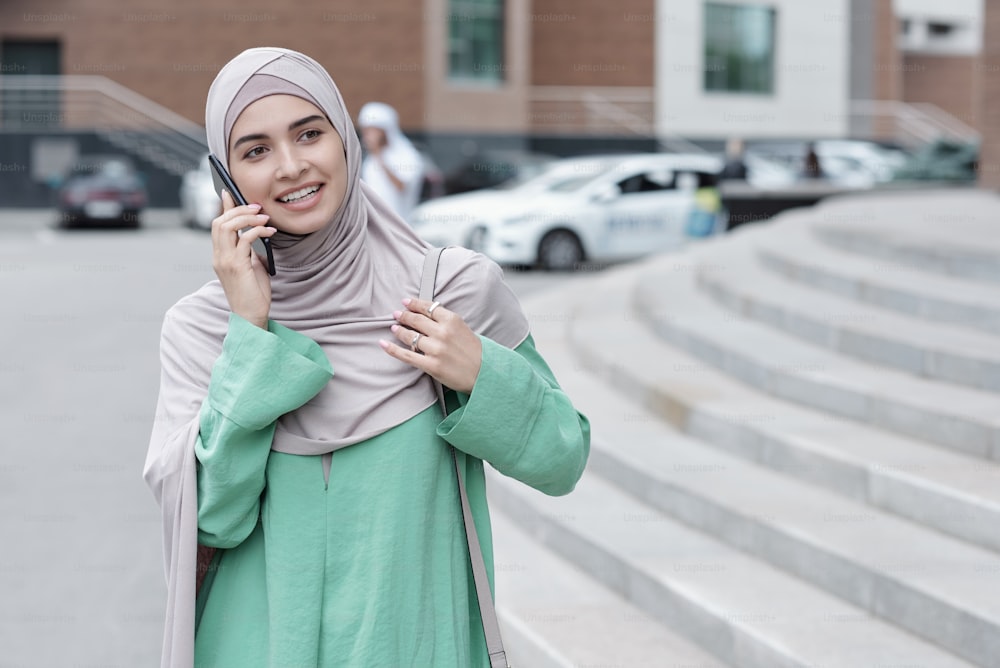 히잡을 쓴 긍정적인 매력적인 젊은 무슬림 여성이 도시 거리에 서서 전화로 채팅을 하고 있다
