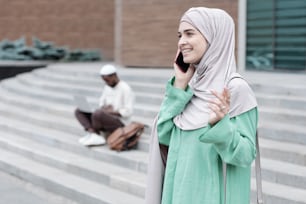 Cheerful attractive young Arabian woman In hijab standing against city building and sharing expressions by phone