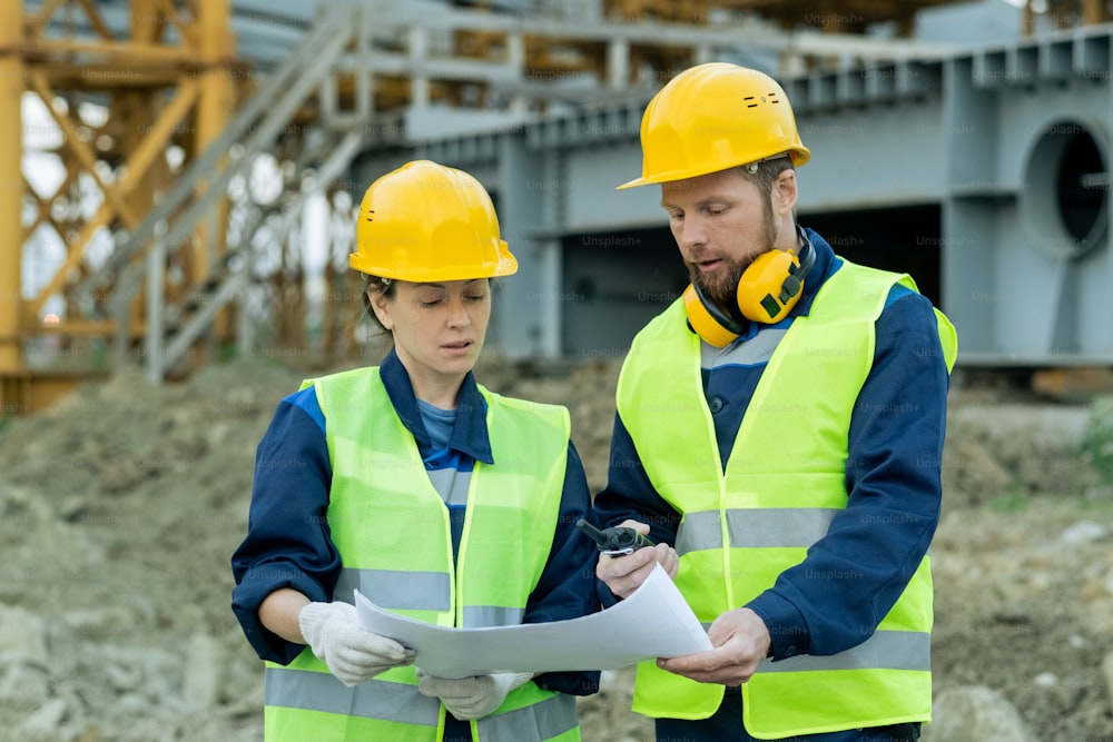 Dos ingenieros en ropa de trabajo y en cascos de trabajo examinando el plano mientras trabajan en equipo en el sitio de construcción