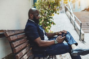 A side view of a mature bald dapper African-American man entrepreneur with a well-groomed black beard, in glasses and blue formal suit, sitting on a street bench and using his smartphone to read news