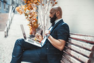 Vista lateral de un hombre de negocios negro maduro maduro, calvo y elegante con una barba bien arreglada, usando su computadora portátil mientras está sentado en un banco de madera de la calle con anteojos en la mano; Hombre africano usando netbook