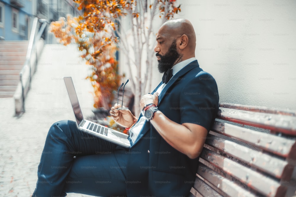 Vue latérale d’un homme d’affaires noir élégant et mûr et mature avec une barbe soignée, utilisant son ordinateur portable assis sur un banc de rue en bois avec des lunettes à la main; Homme africain utilisant un netbook