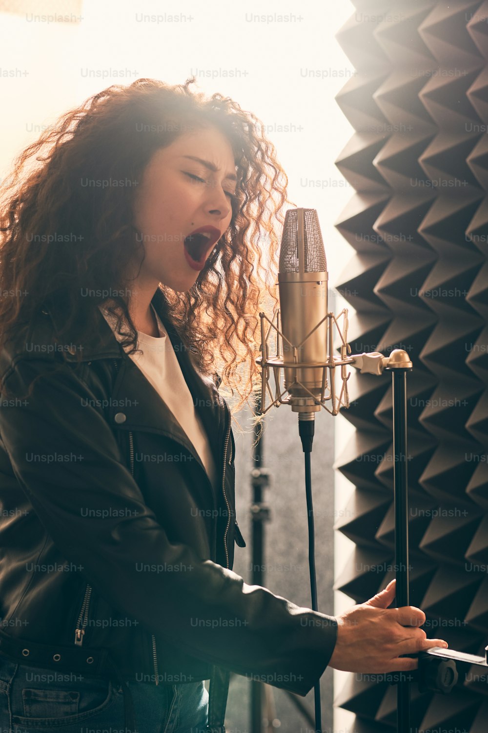Porträt einer schönen lockigen Frau in Lederjacke und Hut, die im Musikstudio auf professionellem Soundequipment Gesang aufnimmt