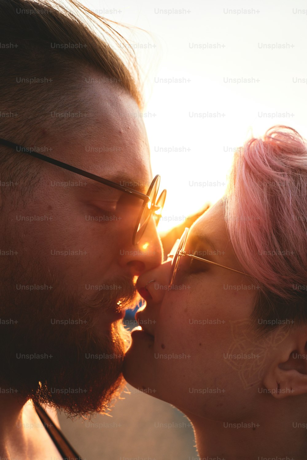Close up retrato de jovem casal em óculos redondos com cabelo colorido beijando ao ar livre na luz natural do pôr do sol. O raio de sol brilha entre eles