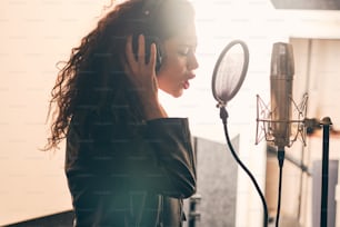Portrait of beautiful curly woman in leather jacket and hat recording vocals in music studio on professional sound equipment, wearing headphones