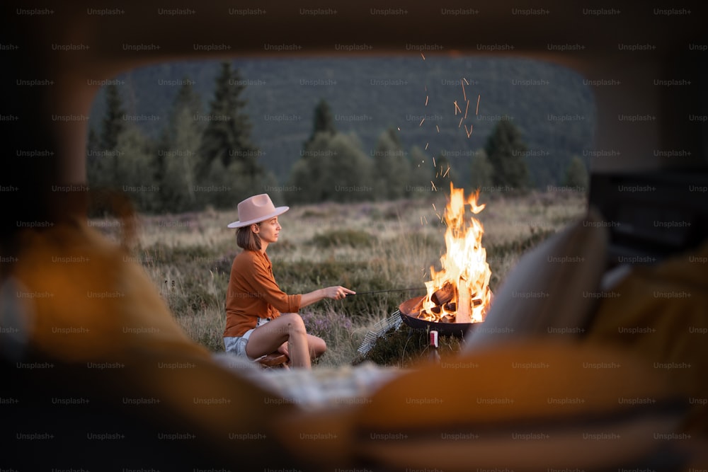 Femme assise près d’un feu de joie lors d’un pique-dans les montagnes, vue de l’intérieur depuis un coffre de voiture. Camping et vacances à la montagne seul, concept de voyage en voiture