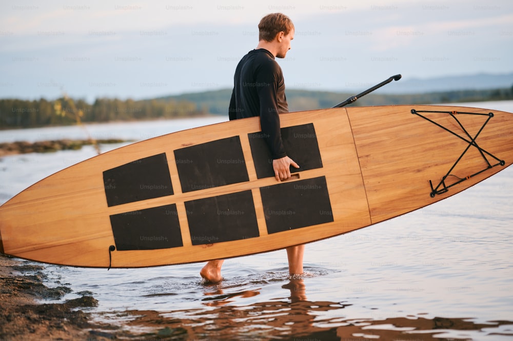 Full body shot of a strong man carrying surf board and a paddle, getting in the water for paddling