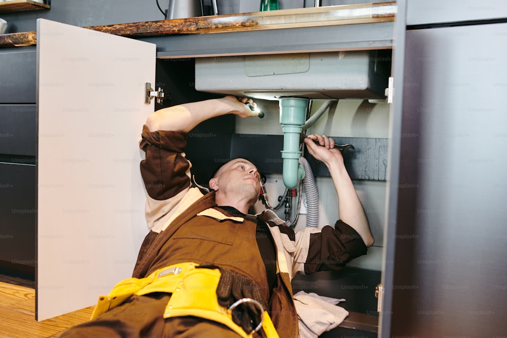 Jeune plombier réparant un nouveau tuyau sous l’évier avec clé