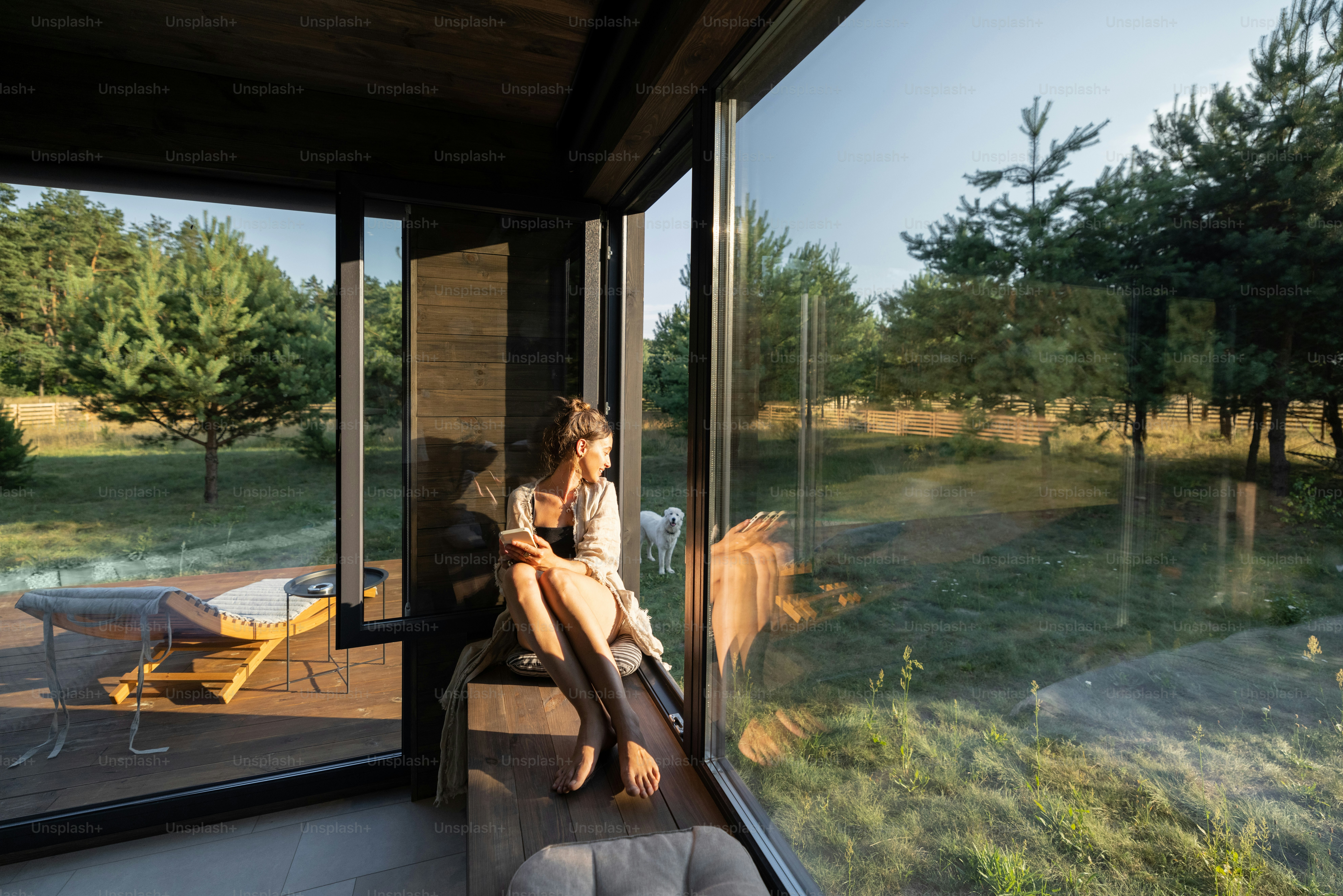 Young woman resting at beautiful country house or hotel, sitting on the window sill enjoying beautiful view on pine forest. Concept of solitude and recreation on nature