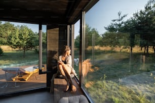 Junge Frau, die sich in einem schönen Landhaus oder Hotel ausruht, auf der Fensterbank sitzt und einen schönen Blick auf den Pinienwald genießt. Konzept der Einsamkeit und Erholung in der Natur