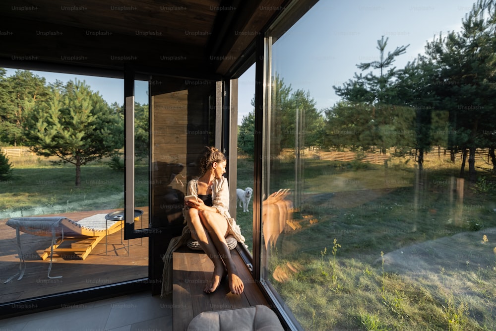 Mujer joven descansando en una hermosa casa de campo u hotel, sentada en el alféizar de la ventana disfrutando de una hermosa vista sobre el bosque de pinos. Concepto de soledad y recreación en la naturaleza
