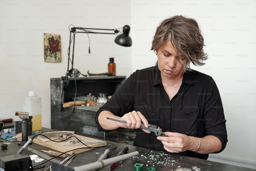 Serious femele jeweler in black shirt sitting at table and measuring blank with caliper while producing ring