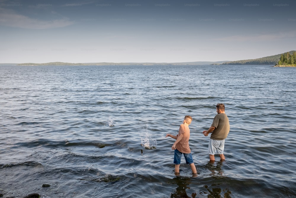 Young man and teenage boy standing in the lake and throwing stones into water on summer day