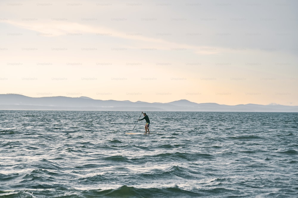 Kleine Figur eines Mannes, der auf einem Surfbrett gegen wilden Natursee paddelt, malerische Aussicht