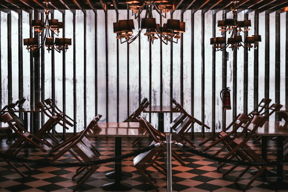 A quarantined indoor restaurant with chairs leaning against the empty tables, checkered floor, and a glass wall; an interior of a closed cafe because of lockdown with three beautiful chandeliers