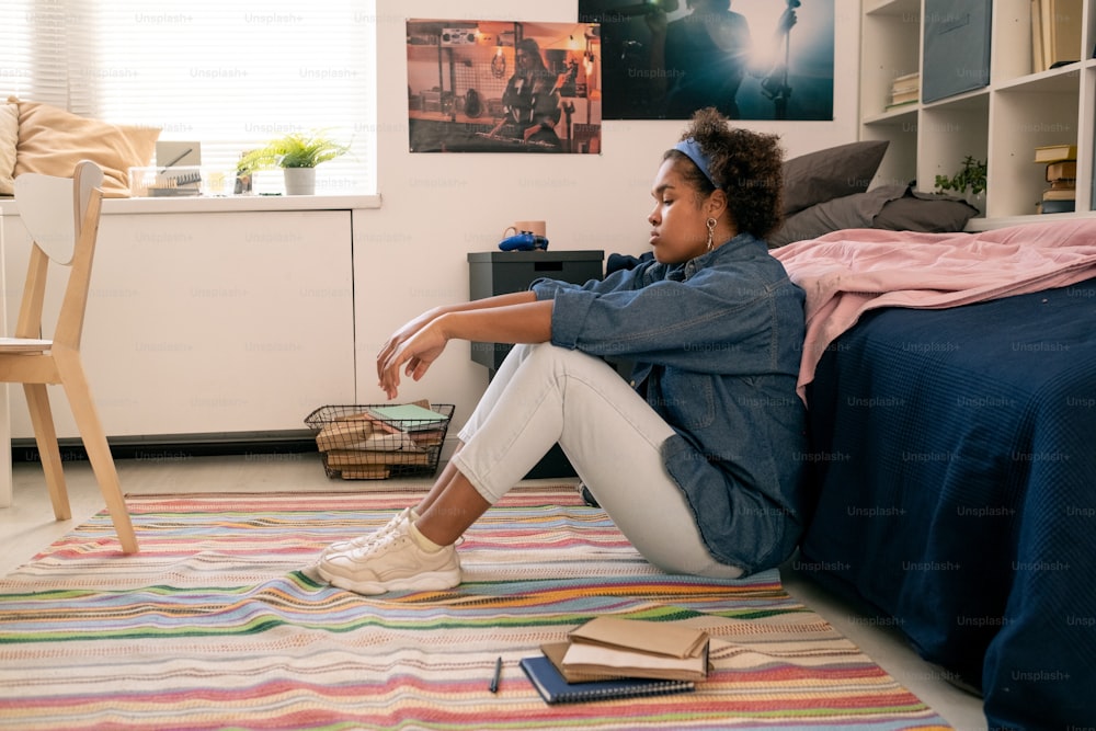 Tired or upset teenage girl in casualwear sitting on the floor by her bed