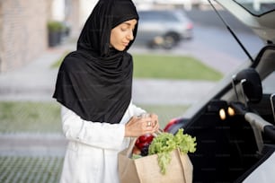 Mulher de negócios muçulmana jovem em hijab indo para casa com mantimentos, de pé com saco de compras na varanda de sua casa