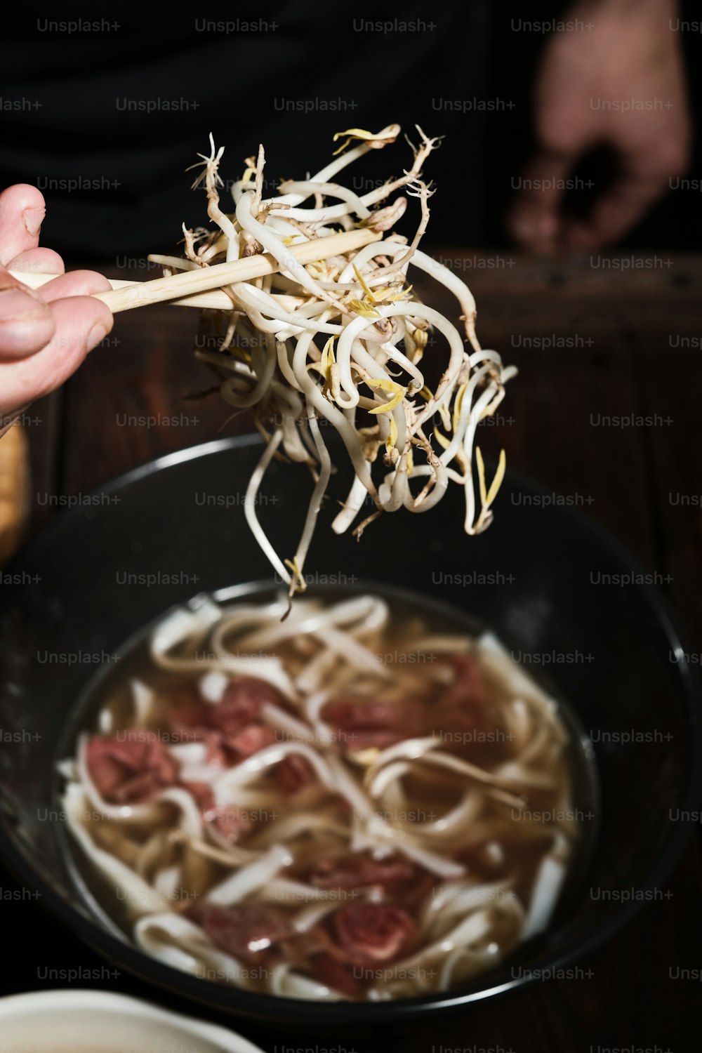 Close up of chef's hands grabbing white bean sprouts, adding them to a bowl of pho bo with beef