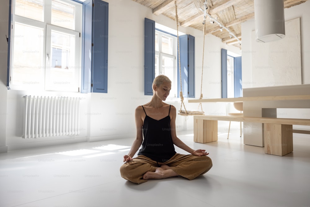 Junge stilvolle Frau, die in einem modernen, hellen Wohnzimmer zu Hause meditiert und Yoga praktiziert. Achtsamkeit, Ruhe im modernen Zuhause