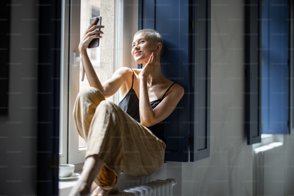 La mujer feliz y elegante se divierte comunicándose por teléfono o haciendo fotos selfie mientras está sentada relajada en el alféizar de la ventana de casa.