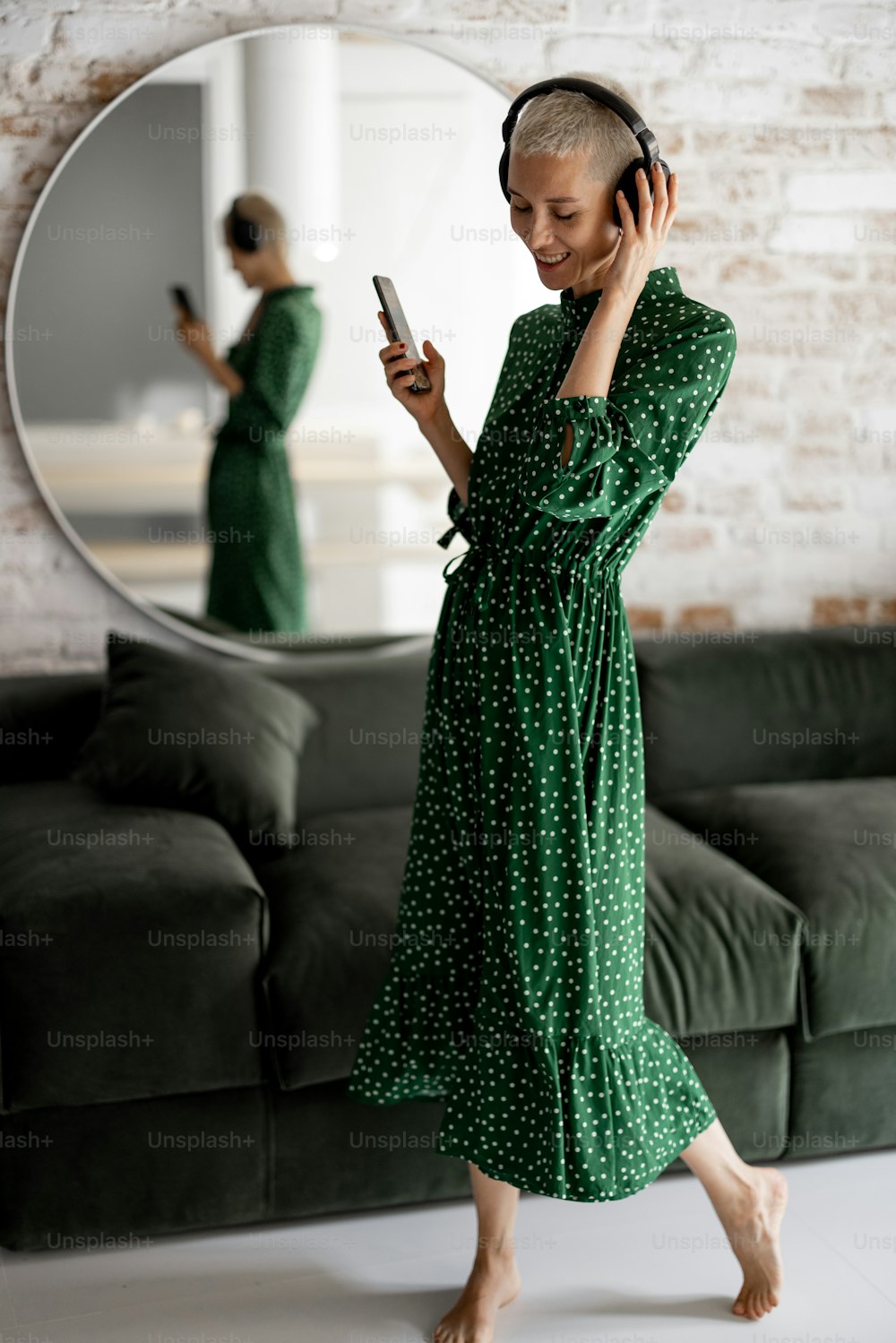 La donna elegante in abito verde gode della musica con le cuffie e il cellulare che balla nel soggiorno di casa
