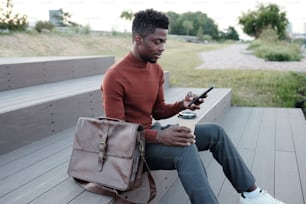 Mobiler afrikanischer Geschäftsmann mit Getränk und Smartphone sitzt auf Treppe im Freien