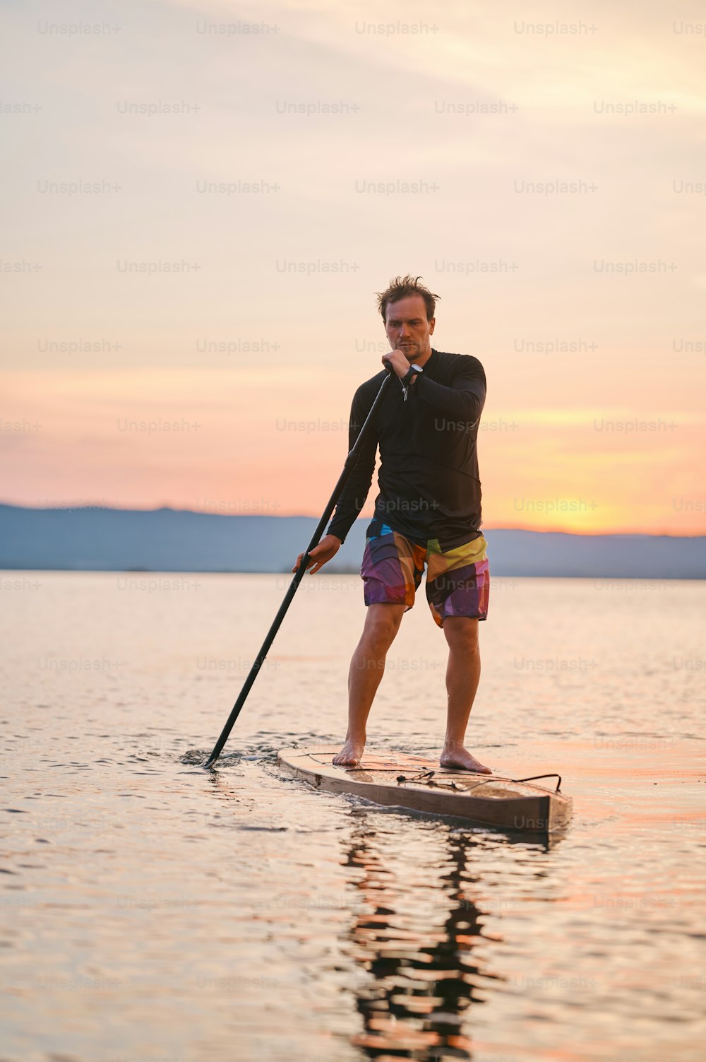 Full body shot of a man with paddle standing on wooden board in quiet water, surfing on sunset with scenic mountain view