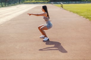 Mädchen und Kniebeuge. Ein Mädchen mit einem schönen Körper, gekleidet in eine Sportgarderobe für eine Übung zur Straffung ihrer Beine und Gesäß auf der Leichtathletikbahn an einem sonnigen Sommertag. Tag für sportliche Aktivitäten