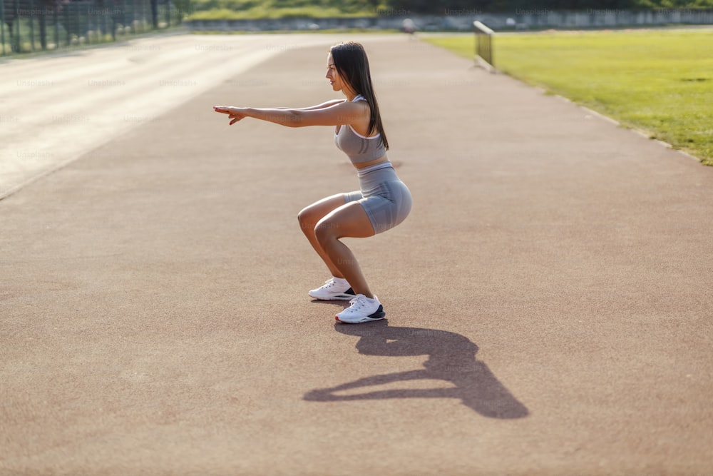 Girl and squat. A girl with a handsome body dressed in a sports wardrobe for an exercise for tightening her legs and buttocks on the athletic track on a sunny summer day. Day for sports activities