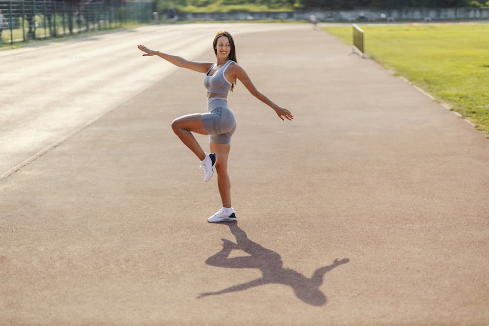 Exercices relaxants et profiter de l’entraînement. Une femme en tenue de sport effectue l’exercice avec des mouvements élégants et féminins. Journée ensoleillée et entraînement au stade d’athlétisme extérieur