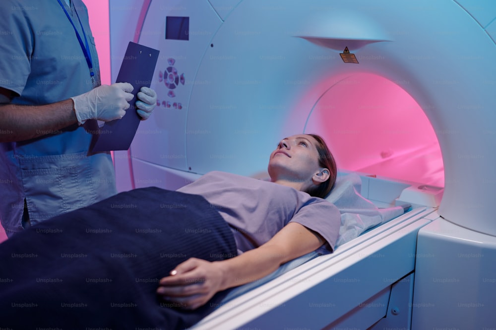 Giovane femmina sdraiata sul lungo tavolo della macchina per la scansione della risonanza magnetica mentre il medico in piedi accanto a lei