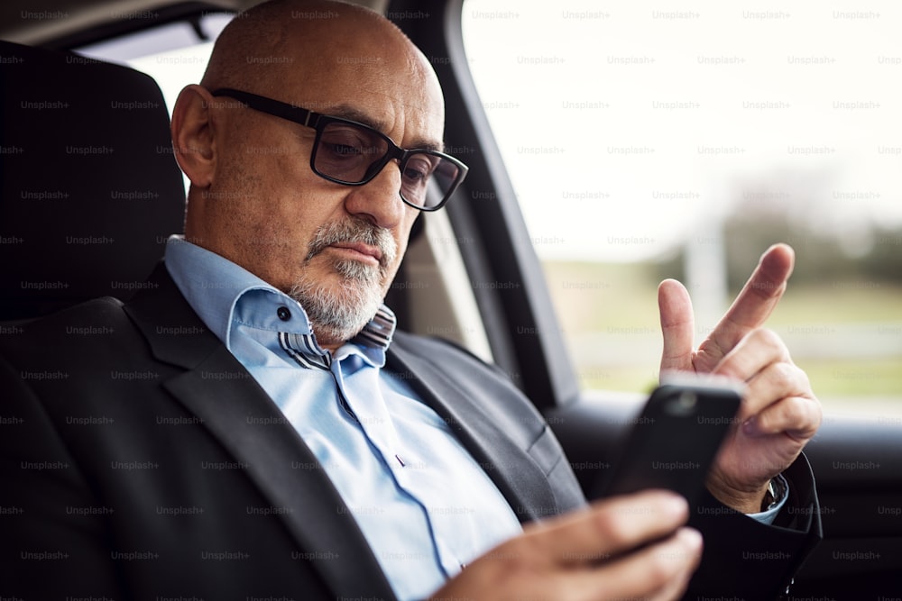 Der reife, ernsthafte Geschäftsmann sitzt in seinem Auto und schaut neugierig in sein Handy.