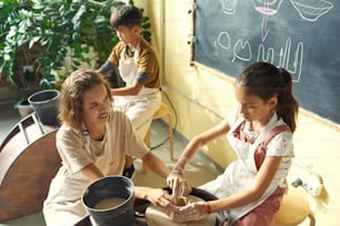 Smiling young Caucasian teacher helping girl in apron to make clay mug using pottery wheel at class