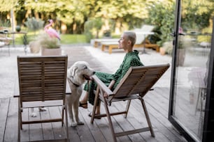 Woman caress dog while sitting on wooden chairs at home terrace. European girl enjoying time wit her pet. Concept of modern lifestyle. Idea of female friendship. Sunny warm daytime